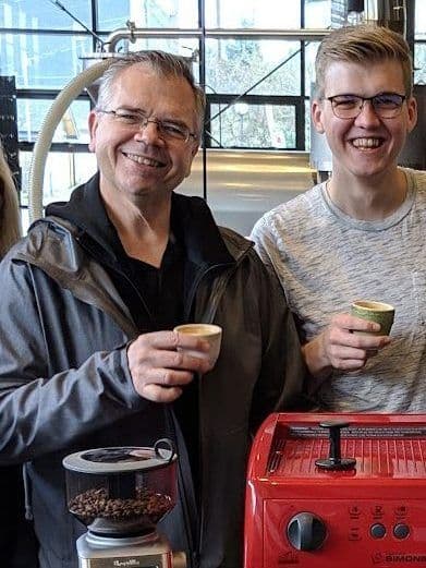Two baristas raise espresso cups beside a red espresso machine and a grinder